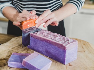 Loaf of purple homemade soap being sliced into bars with orange knife.