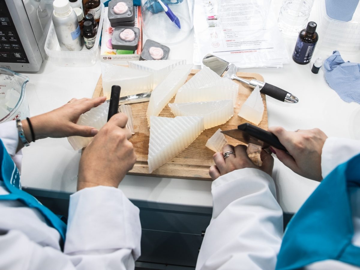 Two people in white coats and blue aprons cutting up a block of solid soap.